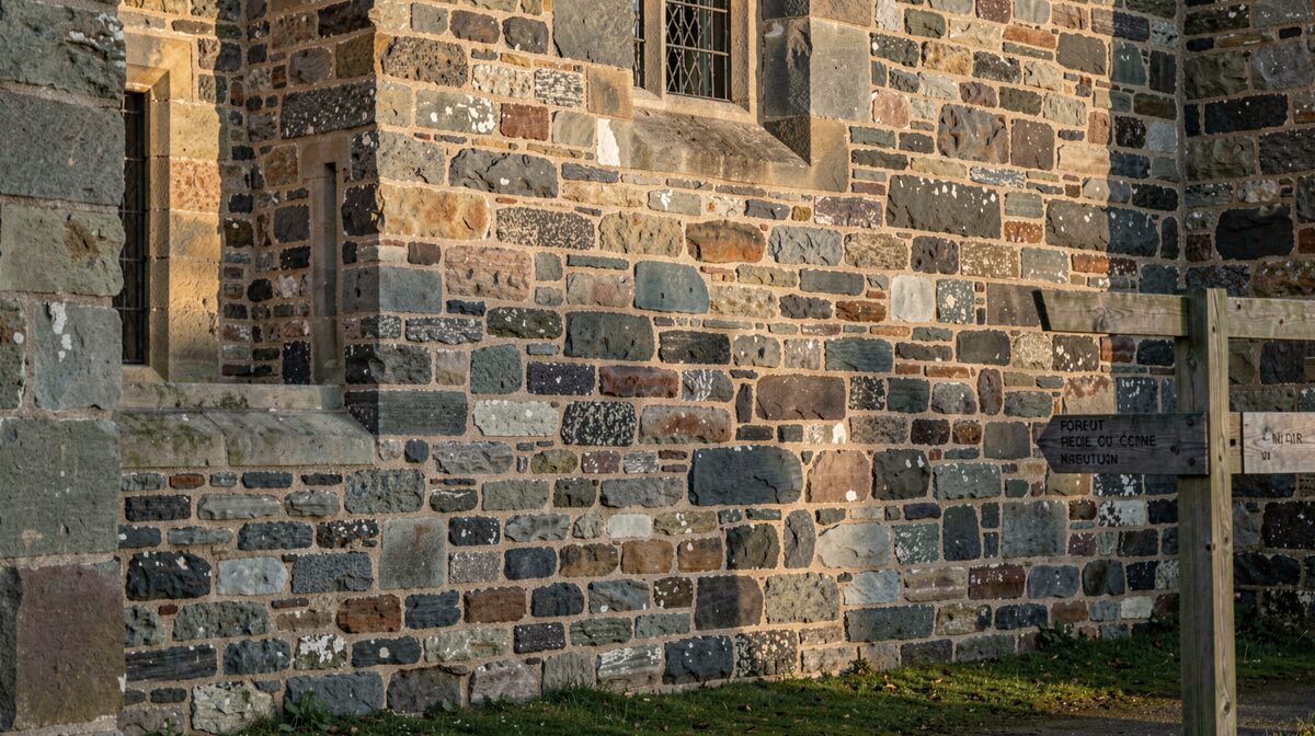 A flint and stone Norman keep wall photographed close up in low morning light, showing mortar lines and window openings, with a footpath signpost at the edge of the frame.